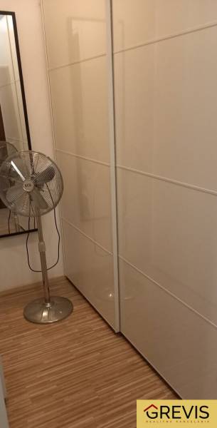A wardrobe in a 2-room apartment with white sliding wardrobes and a standing fan.