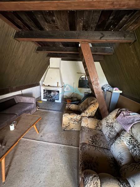 The interior of a cabin with older furniture and a fireplace, wooden beams on the ceiling.