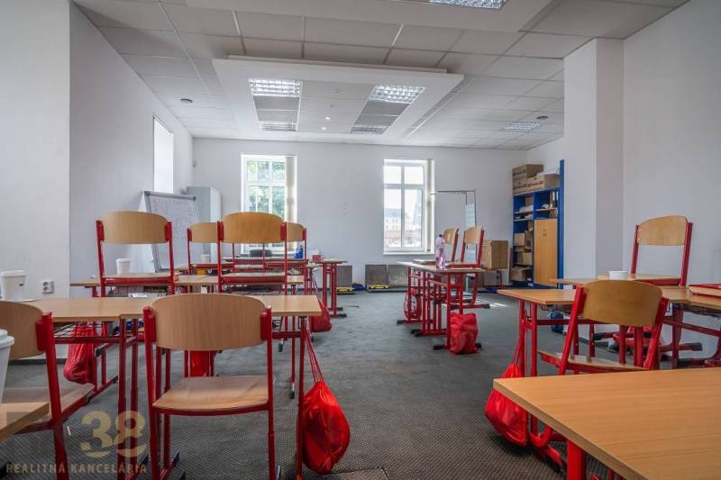 Office in a building with carpeted floor, tables and chairs, cabinets, cardboard boxes.