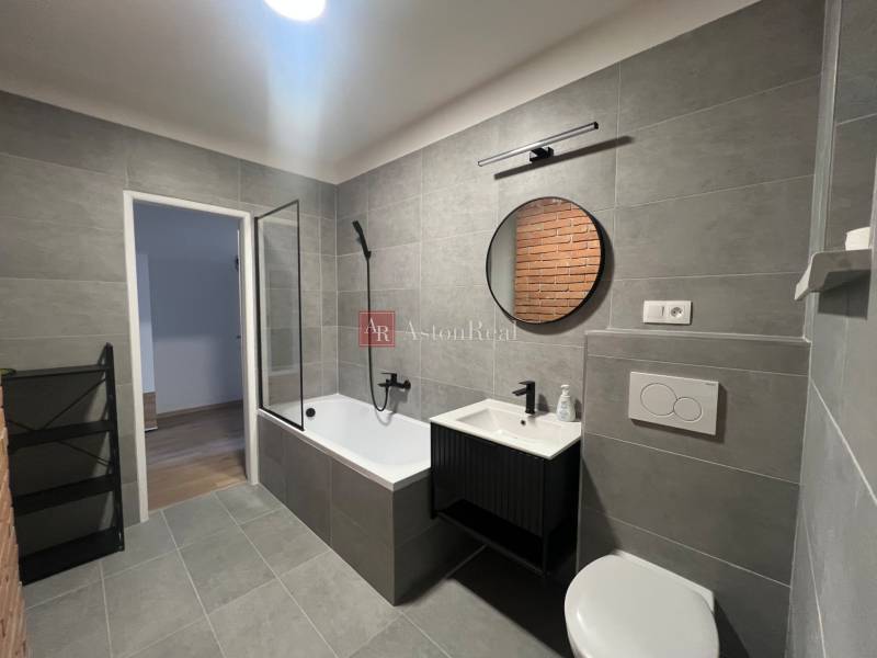 Bathroom in a studio apartment with gray tiles, a bathtub, and a mirror.