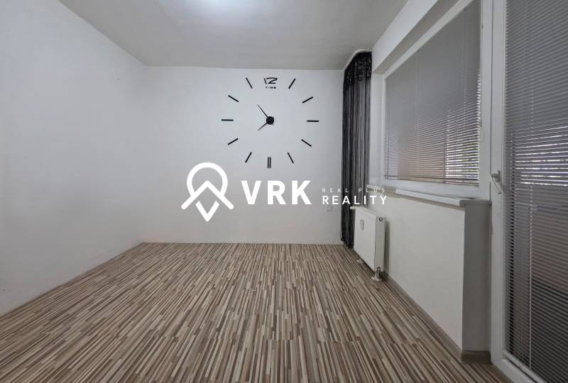 A room in a one-bedroom apartment with a clock on the white wall and a floor with a wooden decor.