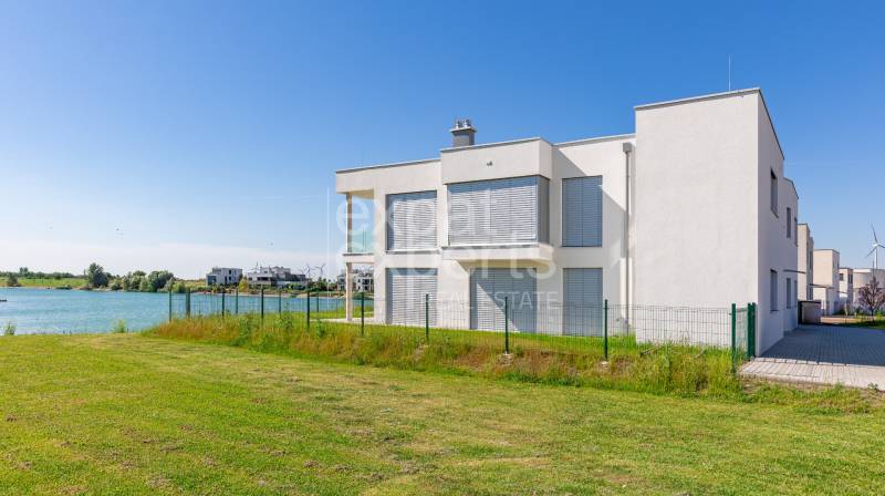 White object by the water, Kittsee, Am Seepark, with a grassy plot and a fence.