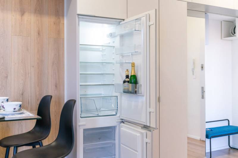 An open fridge with champagne next to a table and chairs in a 2-room apartment with wood-patterned flooring.