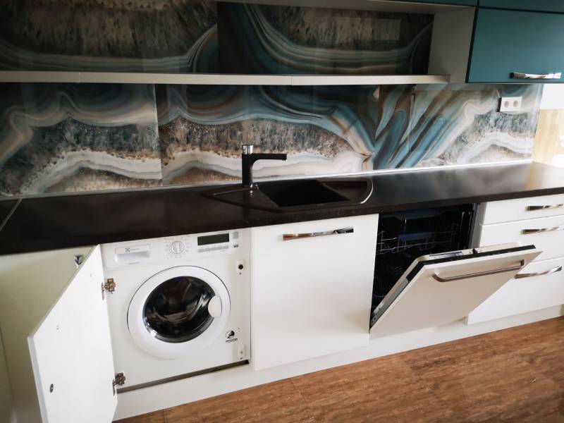 A kitchen with a dishwasher, washing machine, dark countertop, and wood-patterned flooring.
