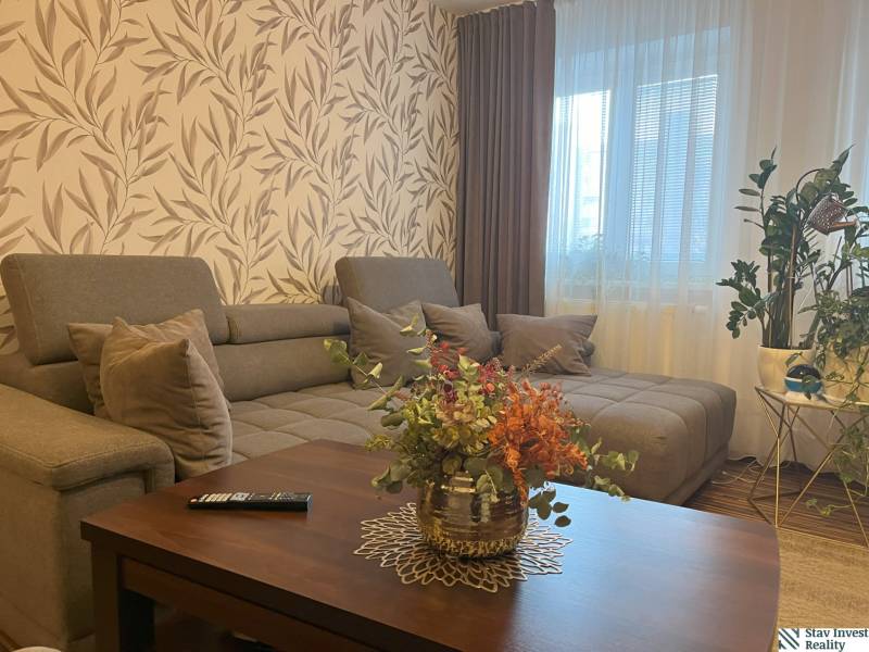 Living room of a two-room apartment with a large sofa, flowers, and patterned wallpaper.