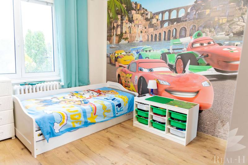 Children's room with car-themed wallpaper, a bed, and storage boxes, wooden decor flooring.