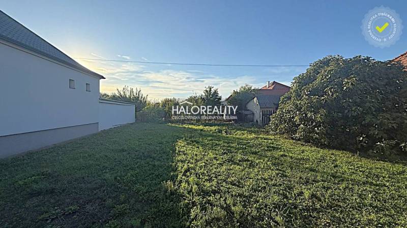 A plot for housing in Kameničná with a grassy area and neighboring houses in the background.