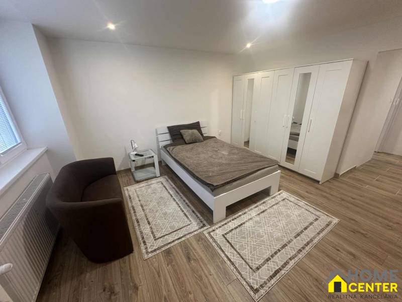 A room with a bed, a chair, and a wardrobe in a studio apartment, with a wooden decor floor.