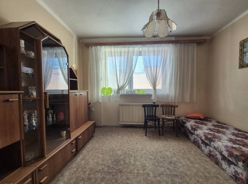 A room in a family house with a display cabinet, bed, chairs, and carpet, illuminated by a window with curtains.