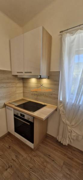 A kitchenette with a cooktop, white cabinets, and a window; flooring with a wood design in a 2-room apartment.
