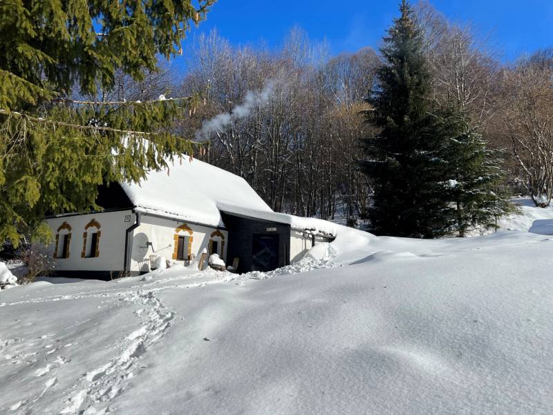A snow-covered cottage in Píla surrounded by winter nature, smoke rising from the chimney.