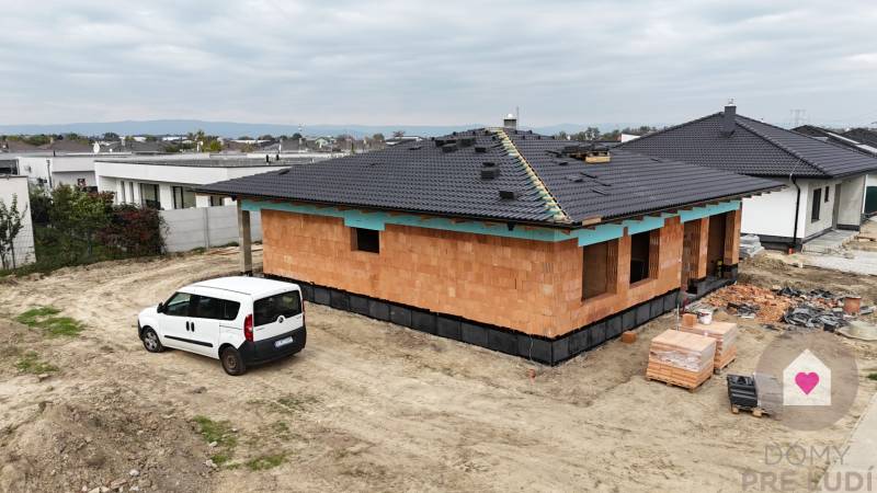 A family house under construction in Bernolákovo, a car in the foreground, neighboring buildings.