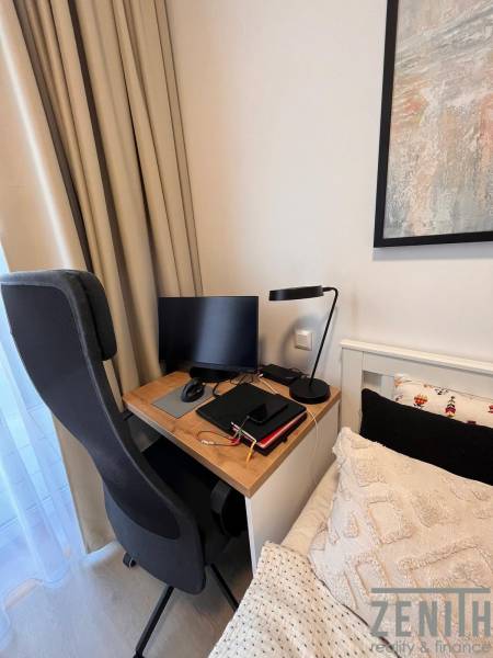 A workspace in a studio apartment with a computer, chair, and lamp next to the bed.