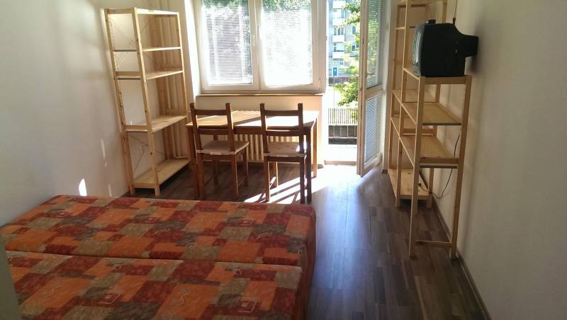 A one-room apartment with a wooden decor floor, a bed, a table, chairs, and shelves.
