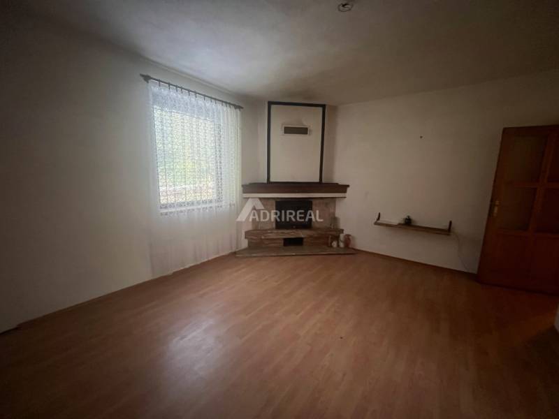 Spacious room in a family house with a fireplace, wooden-patterned flooring, and a large window.