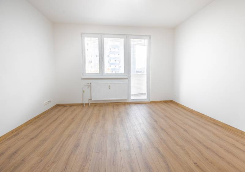 An empty room in a 3-room apartment with a wood-patterned floor and a window.