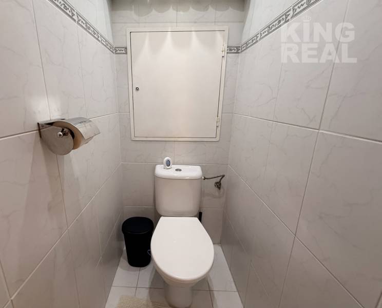 A toilet in a 3-room apartment with white tiles and a toilet paper holder.