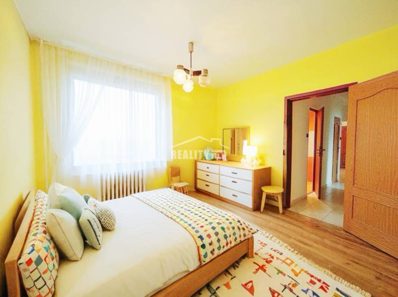 A bright bedroom with yellow walls, a bed, and a wood-patterned floor in a 3-room apartment.