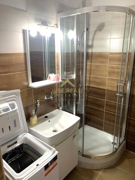 A bathroom in a 2-room apartment with a washing machine, shower, and sink.