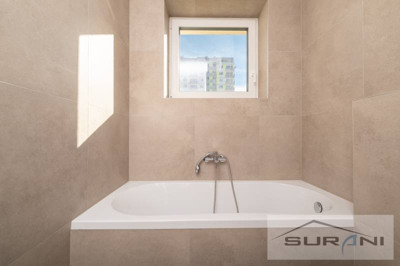 A bathtub with a built-in faucet and a window in a 2-room apartment with warm-toned tiles.