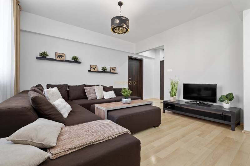 Living room in a two-room apartment with a brown sofa, television, and wooden decor flooring.