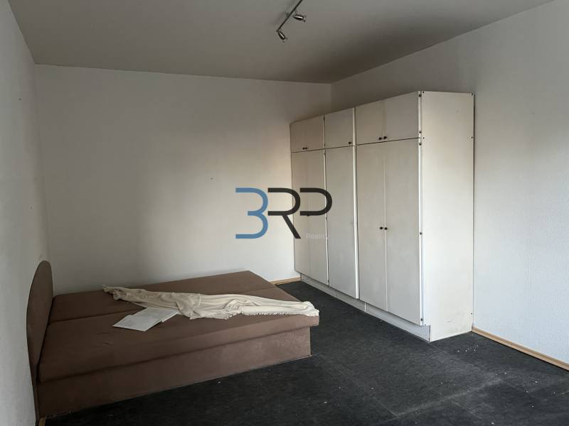 A room in a 2-room apartment with an upholstered bed, a white wardrobe, and a dark floor.