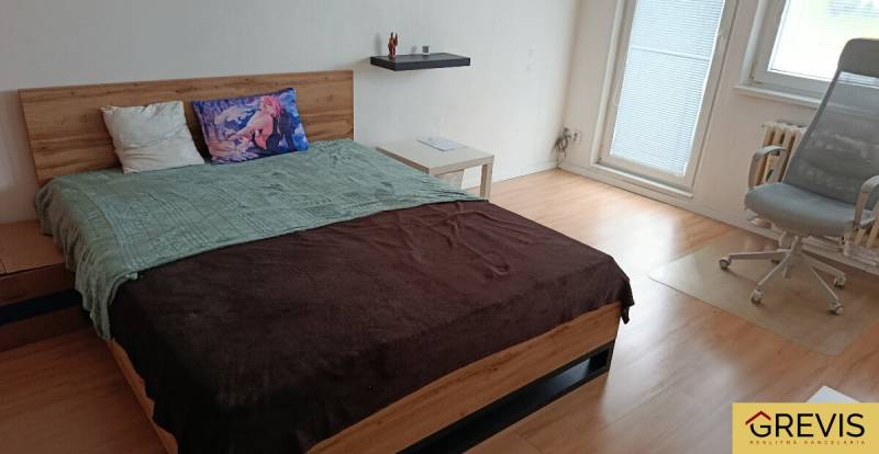 A bedroom in a one-room apartment with a wood-patterned floor, a bed, and an office chair.