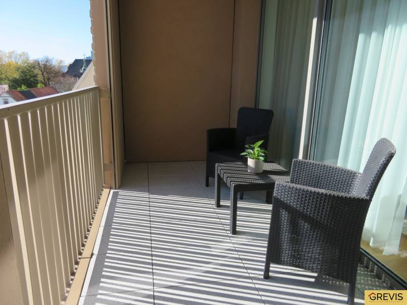 A balcony with rattan furniture in a 3-room apartment, overlooking rooftops and greenery.
