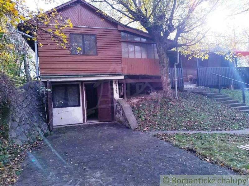 A cottage in Dudince with a stone wall and wooden facade surrounded by trees.