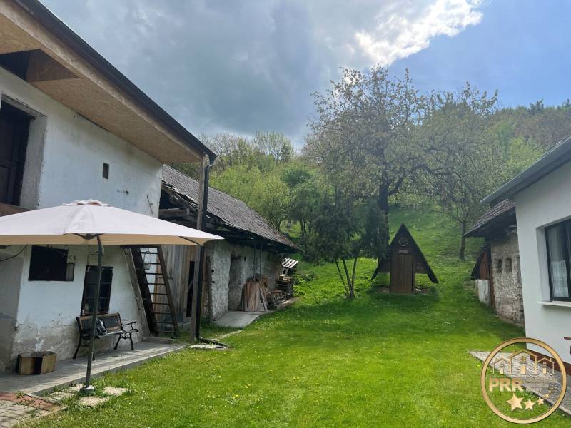 The yard of an older cottage with a gazebo on a bench, a garden in a natural setting on a hill.