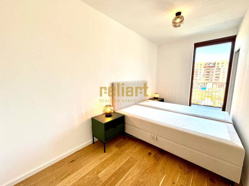 A bright bedroom in a two-room apartment with a wood-patterned floor and a large window.