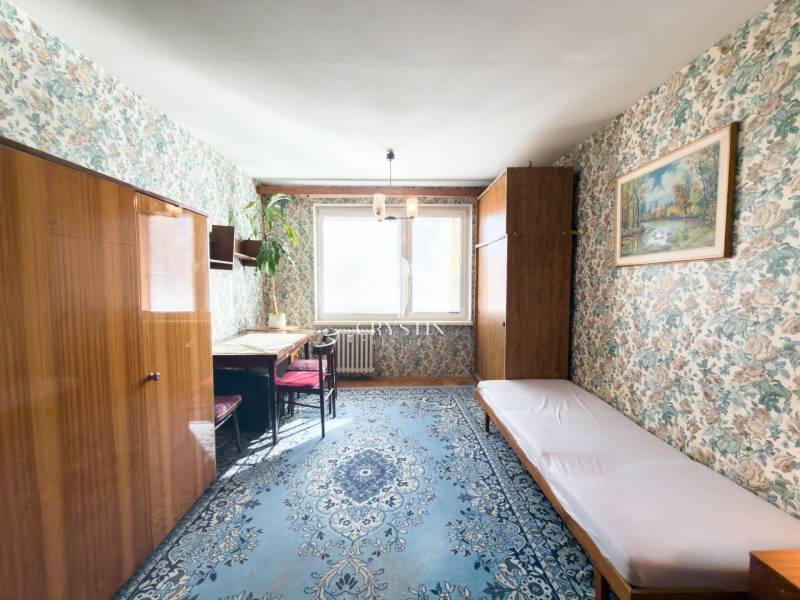 A room in a 3-room apartment with floral wallpaper, a blue carpet, and furniture with a wood finish.