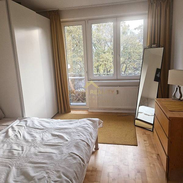 A bedroom in a 2-room apartment with a wood-patterned floor, a bed, and a mirror.