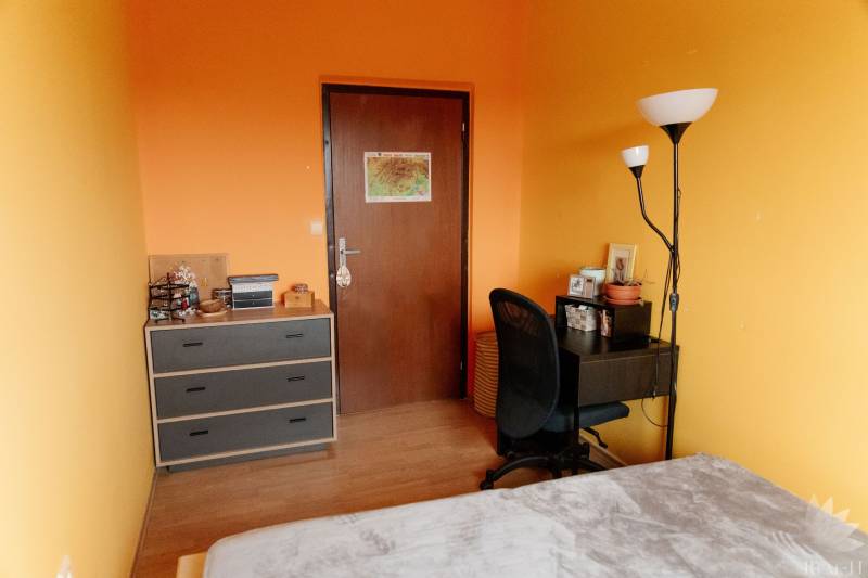 A room in a 2-room apartment with orange walls, a table, and a shelf.