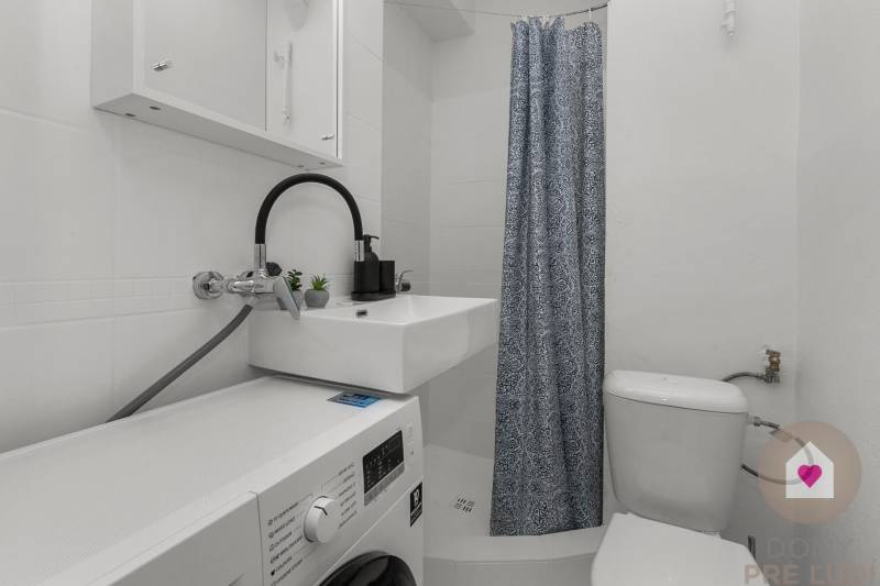 Compact bathroom in a studio apartment with a washing machine and a shower with a patterned curtain.