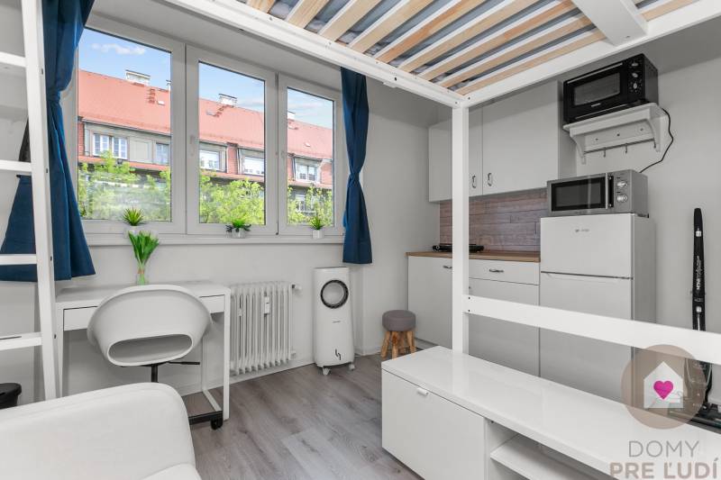 A studio apartment with wood-patterned flooring, a desk, and a kitchen with a microwave.