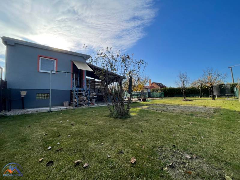 A cottage in the New Settlement in Komárno with a lawn, trees, and a clear blue sky.