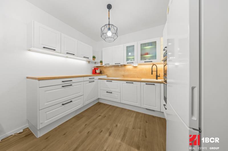A kitchen with white cabinets, wooden countertops, and a wood-patterned floor in a two-room apartment.
