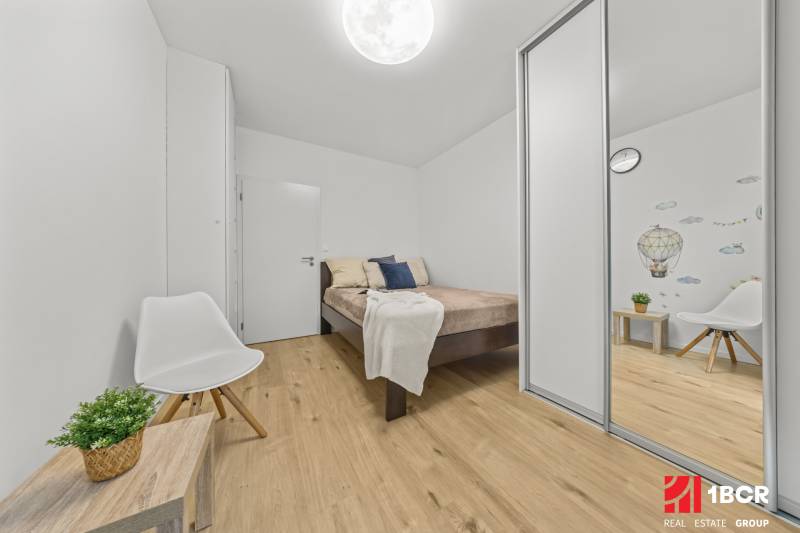 Bedroom in a 2-room apartment with wood-patterned flooring, a white chair, and a wardrobe.
