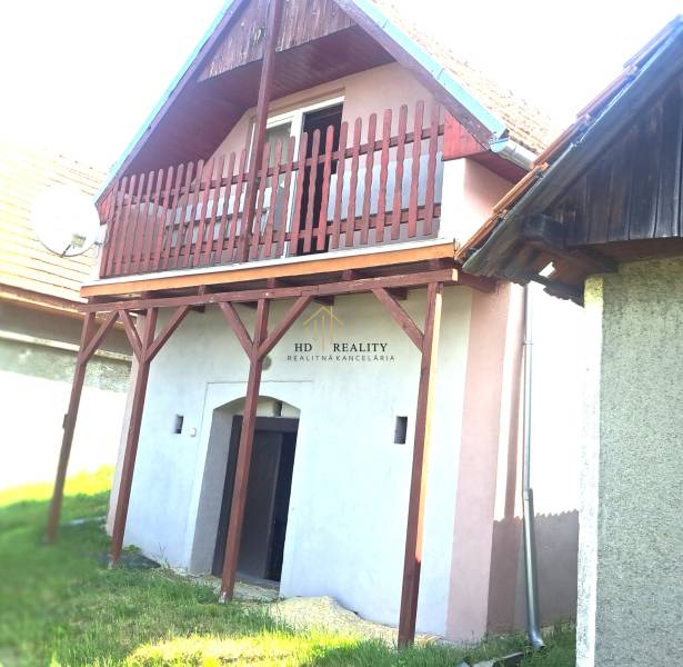 A red balcony on a cottage in Hontianske Trsťany, next to a green lawn and a neighboring house.