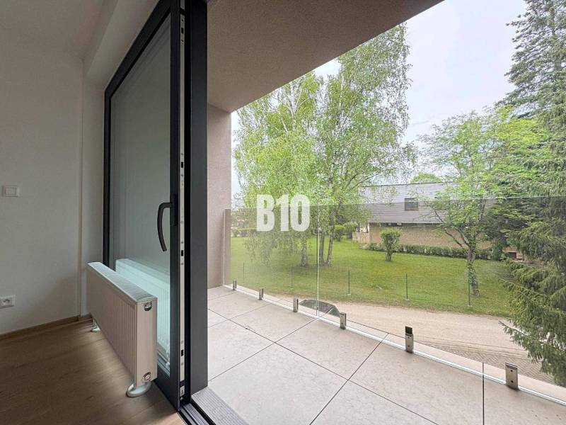 A balcony of a 2-room apartment with a view of greenery and glass railing.