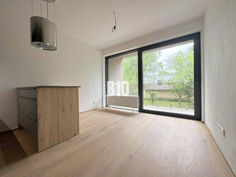 Interior of a 2-room apartment with wooden flooring and a large window overlooking the green exterior.