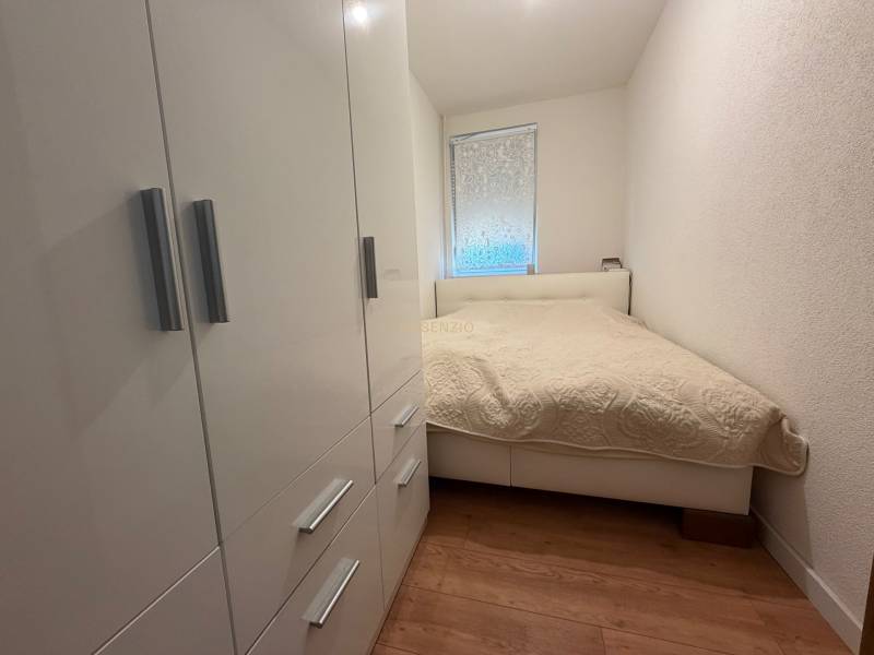 A bedroom in a two-room apartment with a white bed, wardrobe, and wooden decor flooring.