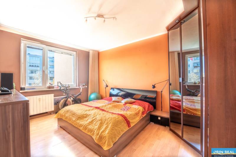 A bedroom with an orange wall, a bed, and a wooden decor floor in a three-room apartment.