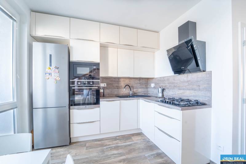 A kitchen in a 3-room apartment with white furniture, built-in appliances, and a wooden decor floor.