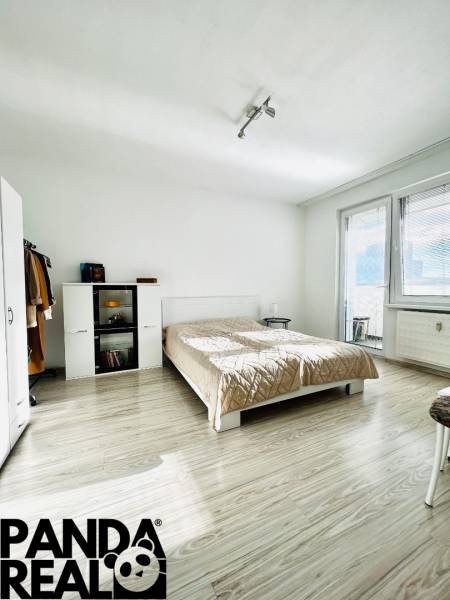 A bedroom in a studio apartment with a wood-patterned floor and light-colored furniture.