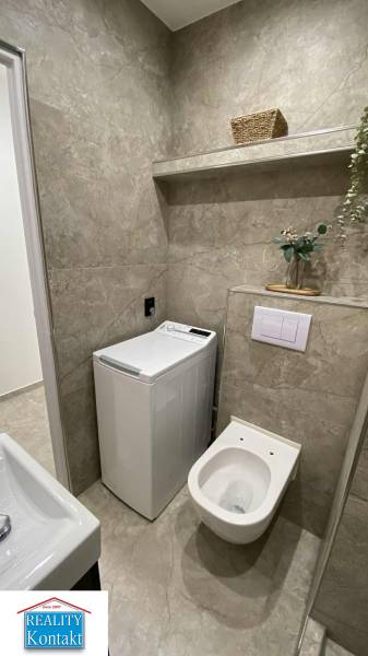 A bathroom in a studio apartment with a washing machine and a toilet, complemented by a storage shelf.