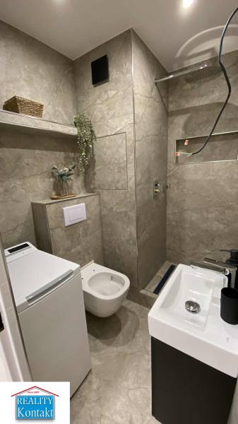 A bathroom in a studio apartment with glossy tiles, a washing machine, and a shower corner.