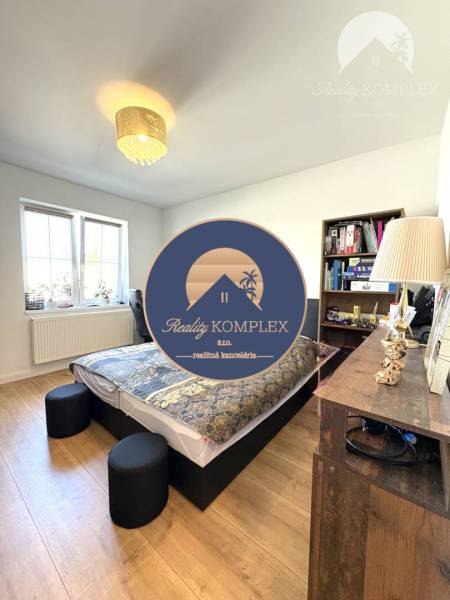 Bedroom in a family house with a bed, wooden decor flooring, window, lamp, and bookshelf.