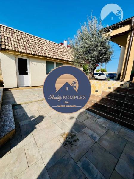 A family house with a sloped roof, a paved yard, and greenery, surrounded by a clear sky.